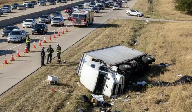 an 18-wheeler rollover on a Texas highway. The truck lies on its side off the road, debris scattered, emergency responders and police securing the area, traffic slowed in the background.