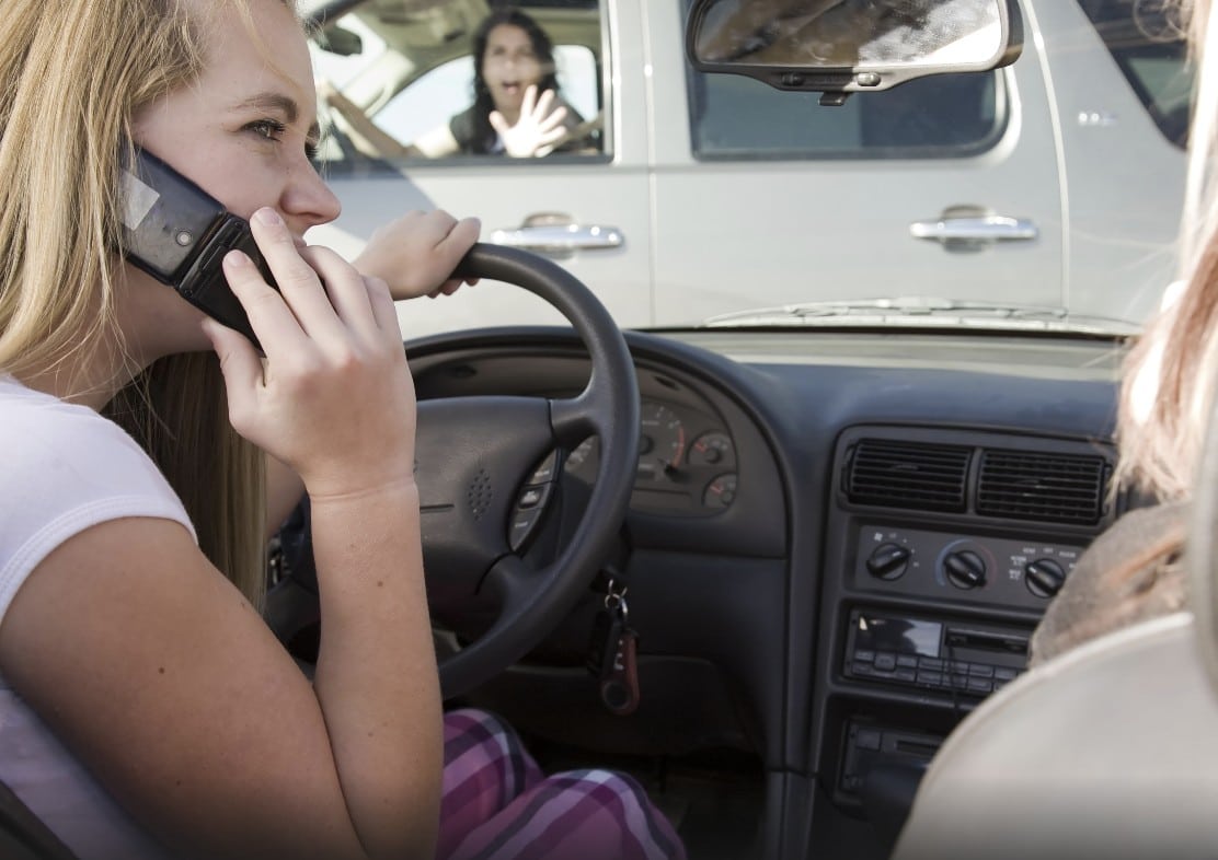 A close-up view from inside a moving car shows a driver speaking on his phone, while the windshield view is blurred, emphasizing the dangerous distraction just before a collision.