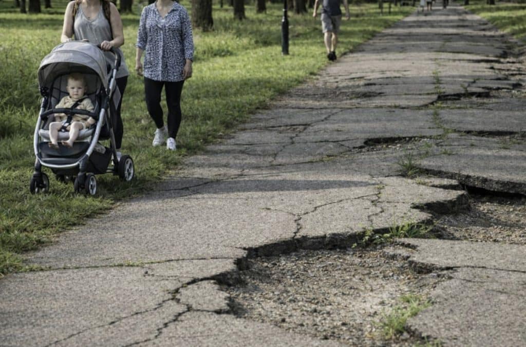 Two women walking with a stroller along a park path with severely cracked and uneven pavement, creating a potential tripping hazard.